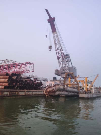 This image depicts an industrial marine scene in Naodoba, Bangladesh, under an overcast sky. The foreground is dominated by dark green water, exhibiting reflections. On the left, a large, flat-top barge carries a dense stack of numerous rusty metal pipes of varying diameters and lengths. Behind these pipes, red metal truss structures are also present on the barge. Centrally, a large white and red lattice boom crane is mounted on a grey floating platform, with multiple hooks suspended from its cables. Adjacent to the crane on the right, a grey and yellow cutterhead dredge vessel is positioned, its circular, toothed cutterhead partially submerged in the water. The dredge vessel features an enclosed cabin structure. No individuals are visible within the frame. The arrangement suggests ongoing or suspended heavy marine construction and dredging activities.