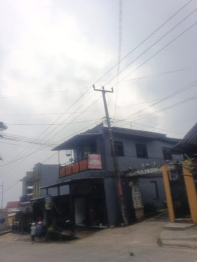 This image depicts a street scene in Dakshin Hichachhara, India, under an overcast sky. A dense network of overhead utility lines crisscrosses the upper frame, supported by a dark utility pole equipped with multiple cross arms and insulators, positioned off-center right.
The central subject is a two-story building, painted dark grey, with a visible pitched roofline. The ground level features storefront openings. The upper level includes a balcony with prominent orange railings, displaying a pink banner and several dark hanging objects, possibly bird cages. To the right of this structure, a stone-faced archway or gate with a partial inscription, "SARNA S," is visible, adjacent to a prominent yellow-painted pillar. To the left, a series of additional multi-story buildings extend into the background.
The foreground consists of a light-colored, unpaved road or pathway. Two indistinct human figures, consistent with children, are present on the left side of the foreground, standing near the base of the central building. Minimal foliage is visible on the far left edge of the frame.