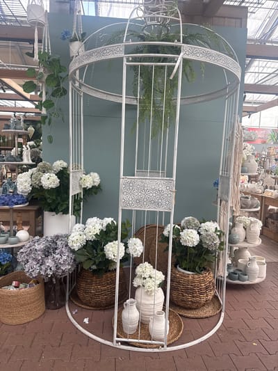 The image depicts a retail display of artificial flowers and decorative pottery in a garden center or flower shop. The primary subject is a large, white, ornate birdcage-like structure serving as a backdrop. Within and around this structure are arrangements of white and purple hydrangeas, some in woven baskets and white ceramic pots. Several white ceramic vases and jugs of varying sizes are placed on woven mats at the base of the display. Hanging plants in macrame holders are suspended from above. In the background, other retail items, including shelves with pottery and decorative objects, are visible. The flooring is made of red brick pavers. The scene is lit by natural light, suggesting an indoor or covered outdoor space. The location is identified as a garden center in Fritzlar, Germany, based on a partial sign visible in the background which includes the word "FRITZLAR". There are no people or animals visible in the image.