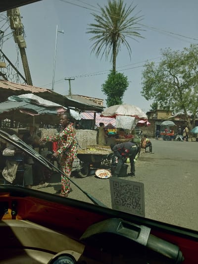 The image depicts an active outdoor market scene, viewed from the interior of a vehicle, likely a tricycle or similar.
**Location:** The specific location of "Kungulu School, Zambia" is not identifiable within the frame. The scene is an urban or semi-urban street market.
**Environment:** The sky is clear and bright, indicating daytime. A paved road occupies the foreground and mid-ground. Buildings, some with metal or red roofs, line the background. Multiple utility poles with electrical wires and transformers are visible on the left, alongside a tall, modern street light pole. A prominent tall palm tree with green foliage stands against the sky in the mid-right, with a smaller bushy tree below it. Another large, leafy green tree is on the far right.
**People:**
- Approximately six to eight individuals are present in varying degrees of clarity.
- In the mid-ground, a person wearing a distinct full-body outfit patterned with red, green, and white geometric shapes stands facing slightly right, positioned near market stalls.
- To the right of this person, another individual in dark clothing (dark shirt, dark pants) is bent over, attending to items on the ground near a wheeled wooden cart.
- Other individuals are partially visible behind market stalls on the left and further down the street on the right, engaged in various market activities such as standing, walking, or tending stalls.
**Subjects/Objects:**
- **Vehicle Interior (Foreground):** A portion of a dark red/orange vehicle dashboard, including a gauge cluster and a black handlebar or control lever, is visible. A black windshield wiper arm extends across the lower-mid portion of the frame. A white square sticker with a QR code and the text "DTLL1424" is affixed to the windshield on the right.
- **Market Stalls:** Several makeshift market stalls are set up along the left and mid-ground, covered by various awnings and umbrellas (grey, red/white striped, plain white).
- **Merchandise:** A wheeled wooden cart, located in the mid-ground center, is laden with produce, possibly green fruits or vegetables. On the ground near the cart, a colorful round platter holds additional items, alongside a dark bucket.
**Actions/Interactions:** Individuals are engaged in typical market activities, including tending stalls, interacting with merchandise, and moving through the area.