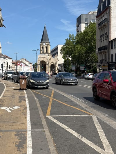 This image captures a street scene in Colombes, France, on a sunny day. The focus is on a street with vehicular and pedestrian traffic, framed by buildings and a prominent church spire. In the foreground, a brown garbage bin on wheels is positioned on a bike lane marked by white lines and a bicycle symbol. Adjacent to the bike lane is a roadway with several cars. A dark grey Peugeot car with license plate "AH-398 GS" is parked near the garbage bin. Further down the street, a silver Renault car is moving in the traffic lane, followed by a red SUV.
The midground reveals a traffic intersection with a traffic light, which appears to be green, indicating that traffic is flowing. Pedestrians are visible walking on the sidewalk. The background is dominated by the architecture of the city, including a church with a clock tower and a steepled roof, flanked by various buildings of different heights and styles. Trees are interspersed among the buildings, adding greenery to the urban landscape. The sky is clear blue with a few wispy clouds. The overall scene depicts a typical day in a French city, with elements of daily life, infrastructure, and historical architecture.