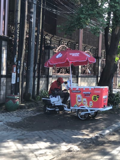 The image depicts a street scene in Buduran, Indonesia, on a sunny day. A vendor wearing a red shirt and cap is seated on a white motorcycle, which is attached to a red ice cream cart. The cart has "Sejuta Jiwa" branding and features images of hands holding cups with thumbs up, along with a large number "8". The cart is positioned on a paved area next to a street, with a decorative patterned fence and a building in the background. To the left of the cart, there is a large, green and red pot. The scene is shaded by a large tree with green leaves. Utility poles with electrical wires are visible behind the fence.