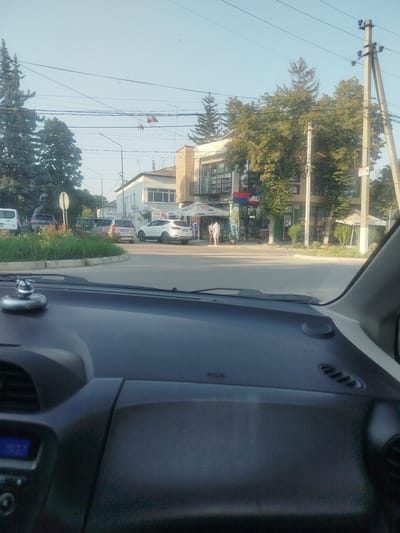 This media file depicts an outdoor scene viewed from inside a vehicle, likely in Criuleni, Moldova. The sun is shining, suggesting it is daytime, possibly late afternoon or early evening, given the soft light. The sky is clear and blue, indicating fair weather.

The scene is a street intersection with several buildings and vehicles. In the foreground, the dashboard of a car is visible, including a digital display showing "9.31". A small, decorative silver object is perched on the dashboard.

Further into the scene, there are several cars parked and in motion. These include a white minivan, a dark SUV, and a silver hatchback. A white SUV is also turning at the intersection. A circular flower bed with red and green foliage is prominent in the middle of the intersection.

Buildings line the street. One building appears to be a commercial establishment with a modern facade, featuring glass windows and balconies, and signage that includes the word "PEPSI" under a blue umbrella. Another building is a white, two-story structure with visible windows and air conditioning units. There are also numerous trees with green foliage, some quite tall and mature, interspersed throughout the landscape and surrounding the buildings. Power lines crisscross the sky, supported by concrete utility poles.

Two people are visible walking outside one of the buildings. They appear to be casually dressed. One is wearing a light-colored shirt and shorts, and the other is wearing a white outfit.

A sign on a utility pole has some visible text, including what appears to be "BATTE" followed by numbers, and another sign in Cyrillic script. The overall impression is a typical street scene in a town or city.