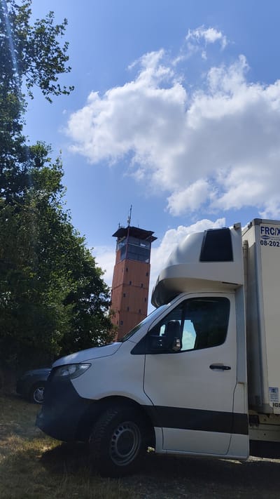 The image captures a bright, sunny day in Hlína, Czechia, with a clear blue sky dotted with fluffy white clouds. In the foreground, the side of a white delivery truck with a silver cab is prominently displayed, taking up the right half of the frame. The truck appears to be parked on a grassy area, with a dark-colored car visible behind it on the left.

Dominating the mid-ground and background is a tall, reddish-brown tower that resembles a lookout or observation tower. It has a distinct structure with multiple levels and windows, topped with antennas and equipment. The tower stands amidst lush green trees, suggesting a rural or semi-rural setting.

The overall atmosphere is serene and depicts a typical outdoor scene on a pleasant day. There are no people visible in the image. Some text is present on the side of the truck, including "FRC/X" and a date "08-2024".