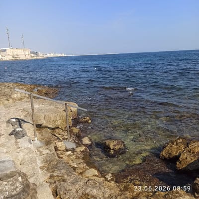 The image captures a view of the Molfetta coastline in Italy, featuring a clear blue sky and calm sea. In the foreground, rugged, light-colored rocks form the coastline, with some areas submerged by the transparent water. A metal railing with two upright supports is visible, offering a pathway or support structure along the rocky edge. The water is shallow near the shore, allowing the seabed and rocks beneath the surface to be seen. In the distance, a cityscape stretches along the horizon, characterized by a line of buildings and a prominent, modern-looking structure with two tall antennas. The presence of a white floating object in the water suggests a marker or buoy. The timestamp "23.06.2026 08:59" is overlaid on the bottom right of the image.