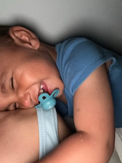 A close-up, over-the-shoulder shot captures a young child with their eyes closed, resting their head on a person's shoulder. The child is wearing a blue t-shirt and has a light blue pacifier with a bunny-like design in their mouth. The child's right ear is visible in the upper left corner of the frame. The background is a plain, light gray surface, suggesting an indoor environment. The lighting appears to be dim, with light illuminating the child's face and arm. The image is cropped closely, focusing on the child's head and upper body, and the shoulder they are resting on. No specific location in Rakitovo, Bulgaria, can be determined from the visual information provided.