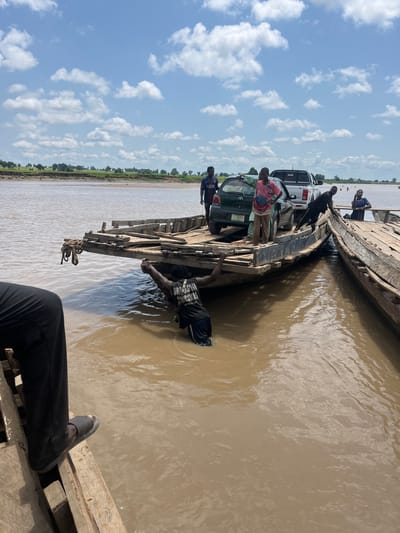 An outdoor, medium shot captures a ferry operation on a wide, muddy river under a cloudy sky in Nemnei, Nigeria. Two wooden barges, tied together, are positioned side-by-side. A green sedan is loaded onto the left barge, while a white pickup truck is on the right barge. Several individuals are present. On the left barge, a person is partially submerged in the water, reaching towards the edge. Another person, seen from the waist down, stands on the wooden planks of the left barge. On the right barge, two people are standing near the pickup truck, and another person is seated. In the distance, the riverbank is lined with green vegetation and trees. The water appears calm with ripples.