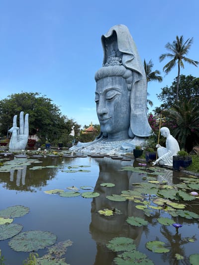 This image depicts a serene outdoor scene featuring a large, serene statue of a deity, possibly Guan Yin, with a serene expression. The statue is made of a pale, weathered material, possibly stone or concrete, and appears to be partially submerged in a pond filled with lily pads and a few blooming water lilies, some of which are purple. To the right of the main statue, a smaller white statue of a person kneeling and holding a staff can be seen. In the foreground, a hand from the main statue emerges from the water, with three fingers extended upwards. The background shows lush green trees, including palm trees, and a clear blue sky. The overall ambiance suggests a tranquil, spiritual setting, likely a temple garden or a place of worship. The lighting indicates it is daytime, possibly late afternoon given the soft light and reflections on the water. The location is identified as Hòa Đa, Vietnam.