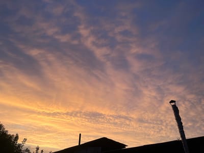 The image depicts a dramatic sunset sky over a silhouetted landscape. The sky is filled with a variety of clouds, from wispy, elongated formations in shades of orange and yellow near the horizon to smaller, more textured clouds in purples and blues higher up. The lower part of the image shows the dark silhouettes of rooftops and trees. A tall, cylindrical object, possibly a chimney, stands out prominently on the right side, wrapped in a textured material. The overall impression is one of a serene and beautiful end to the day. The time of day is clearly sunset, characterized by the vibrant colors in the sky. The weather appears clear with a dramatic cloud formation. The location cues, such as the style of roofing and the presence of the chimney, are consistent with a residential or industrial area in Kazakhstan. The emotions evoked are peacefulness and awe at the natural beauty of the sunset. There is no visible text in the image.