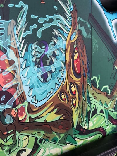 This is a close-up photograph of a printed image, possibly a mousepad or poster, featuring a vibrant, cartoonish depiction of a monstrous, alien creature in a surreal, green-hued environment. The creature's design is complex and grotesque, with multiple bulbous eyes featuring red and yellow irises, sharp teeth, and tendrils. It is surrounded by swirling blue watery elements and green, glowing energy lines. The overall aesthetic suggests a science fiction or fantasy theme. The image is not located in Mirnoye, Ukraine, as it is a manufactured product and not a photograph of a real-world scene.