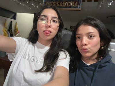 Two women are posing for a selfie in Vic's Cantina, El Oasis Valsequillo, Mexico. The woman on the left is wearing a dark blue hoodie and gold earrings. The woman on the right is wearing glasses and a white t-shirt. There are glasses and bottles behind them.