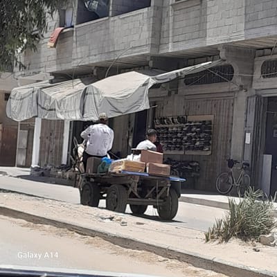 In the city of Nuseirat, Palestinian Territories, a man wearing a white shirt and a cap is seated on a horse-drawn cart. He appears to be operating the cart. Another man, wearing a light-colored shirt and a cap, is seated on the cart as well, facing away from the camera. The cart is loaded with several cardboard boxes and a blue bag. The cart is moving along a street. In the background, there is a building with a covered awning over a storefront. The storefront displays shelves of shoes. A bicycle is parked to the right of the storefront. The ground is a dirt and paved surface.