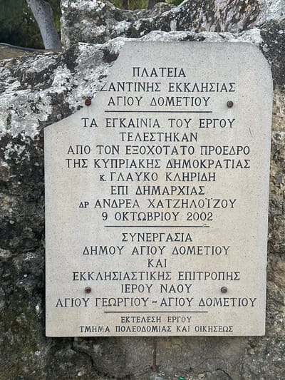 The image displays a light-colored, rectangular stone plaque with rounded corners, firmly embedded into a rough, dark grey rock formation. The plaque is angled slightly, with its text presented vertically. Two metallic bolts are visible on the left side of the plaque, securing it to the rock. The rock itself shows significant natural texture, including crevices and patches of light-colored lichen or moss-like growth, suggesting an outdoor setting. Hints of blurred greenery are visible in the background.

The Greek text on the plaque commemorates the inauguration of a project in Agios Dometios, Cyprus. It reads:
"PLAZA OF ZANTINIS CHURCH, AGIOU DOMETIOU
THE INAUGURATION OF THE PROJECT TOOK PLACE
BY HIS EXCELLENCY THE PRESIDENT OF THE REPUBLIC OF CYPRUS, MR. GLAFKOS CLERIDES
DURING THE MAYORALTY OF DR. ANDREAS HATZILOIZOU
OCTOBER 9, 2002
COOPERATION:
MUNICIPALITY OF AGIOU DOMETIOU
AND
ECCLESIASTICAL COMMITTEE OF THE HOLY TEMPLE OF AGIOU GEORGIOU - AGIOU DOMETIOU
PROJECT EXECUTION:
DEPARTMENT OF URBAN PLANNING AND HOUSING"

The plaque thus serves as a historical marker, documenting the opening of a plaza on October 9, 2002, under the patronage of the then-President of Cyprus, and the collaborative efforts of local authorities and religious institutions.