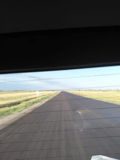 The image shows a view through a car window of a straight, asphalt road stretching into the distance. The road is bordered by a dry, grassy field on the left and a lighter colored verge on the right. The sky is clear and blue with some scattered clouds. There are thin black lines across the image, indicating the defroster lines of the car window. A white sign is visible in the field on the left side of the road. The scene appears to be a rural or roadside environment. The location is identified as Молодёжный, Kazakhstan.