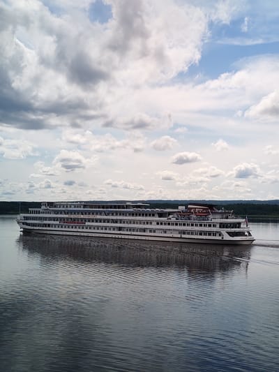 A large white passenger ship, identified by the Cyrillic lettering "ВИССАРИОН БЕЛИНСКИЙ" on its side, navigates a calm body of water, likely a river, under a cloudy sky. The ship has multiple decks, indicated by rows of windows. Its reflection is clearly visible on the water's surface, creating a symmetrical image. In the background, a dense line of dark green trees forms the distant shore, meeting a pale blue sky dotted with white and gray clouds. The overall scene suggests a serene, perhaps summer day, with the ship moving away from the viewer towards the right side of the frame. The specific location is the city of Shchetinino, Russia.