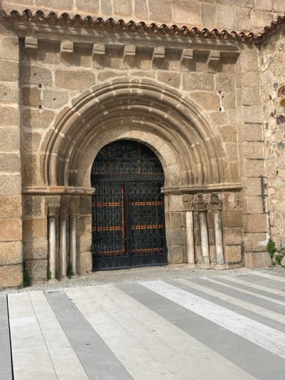 This image presents a close-up view of the exterior wall of a historic building in Mérida, Spain, bathed in bright daylight. The primary focus is an elaborate, partially visible circular window set into a robust stone wall. This stone wall is constructed from irregular, light brown and grey blocks, exhibiting a weathered texture.
The circular window, characteristic of a rose window, is framed by multiple layers of carved stone molding, creating depth and architectural detail. Within its lower half, a dark, intricate iron grill with a repeating geometric pattern secures the opening. Directly beneath this, a rectangular window, also protected by a matching dark iron grill, is visible.
To the left of the main stone wall, a decorative vertical element is present, composed of a series of terracotta tiles arranged in a wavy, scalloped pattern, suggesting an ornate architectural feature or roofline. On the far right, a contrasting modern wall with distinct horizontal stripes of alternating light and dark grey materials, possibly stone or concrete, stands adjacent to the older structure. Small tufts of green vegetation sprout from cracks in the ancient stonework. The lighting indicates a sunny day, casting subtle shadows that highlight the textures of the stone. No people or activity are depicted, and no visible text is present.