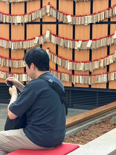 In Ota, Japan, a man is seated, facing away from the camera, looking down at his phone. He wears a dark blue short-sleeved shirt and light beige pants. A black bag is slung across his back. Behind him, a wall is covered with many small wooden plaques, each tied with red string and hanging in rows. These plaques appear to be prayer or wish tablets. The environment suggests an outdoor or semi-outdoor setting with a wooden bench and some greenery visible in the foreground.