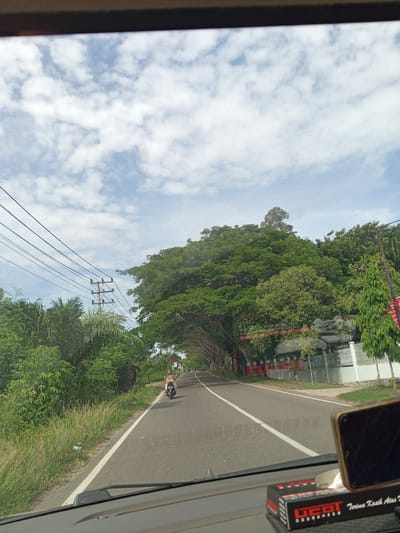 Captured from inside a vehicle, the image shows a road in Pulorungkom, Indonesia. A motorcycle is traveling along the road. Vegetation lines the roadside, including a canopy of trees, along with overhead electrical wires. On the right side of the road is a red and white building. The sky is blue with scattered clouds. A tissue box and a phone are visible on the dashboard of the vehicle.
