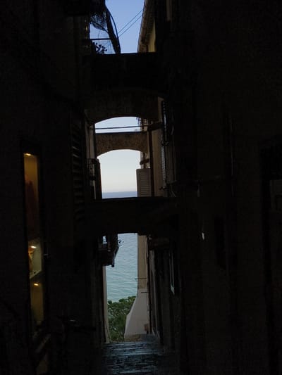 A vertical, low-angle shot captures a narrow street in Sperlonga, Italy, leading towards the sea. The buildings on either side are dark, creating a tunnel-like effect. Glimpses of the Mediterranean Sea are visible through arched openings between buildings. The upper portion of the image shows a clear blue sky. The street is paved with stones, and there is a tree on the right side of the image. The scene is dimly lit, suggesting it is either early morning or late evening.