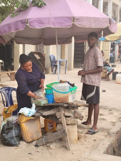 The image captures an outdoor street vending scene in Ifite Awka, Nigeria, under what appears to be warm, daytime conditions, shaded by a large, faded purple umbrella. The ground is sandy.
Two main individuals are visible. In the foreground, a woman wearing a dark long-sleeved top and skirt stands beside a makeshift stall, constructed from stones, wood planks, and possibly concrete blocks. She is actively engaged in handling items, holding a clear plastic bag filled with green produce, and looking down at her work. Her expression appears focused. Adjacent to her on the ground is a blue plastic chair.
Further back and to the right, a young man stands upright, wearing a light plaid short-sleeved shirt and dark shorts. He is looking towards the camera with a neutral to slightly smiling expression. He wears dark sandals on his feet. In the distant background, another person is lying down, partially obscured.
The stall itself holds various containers. On top, there are clear plastic jugs or bowls, some containing brownish liquids, and a blue bucket. Below the main stall structure, a large yellow jerrycan and a smaller yellow plastic container are visible. Several plastic bags are scattered around the stall and on the ground. One white plastic bag near the woman clearly shows part of the brand name "INDOMIE". Another bag on the ground contains yellow granular material, possibly food items. The overall setup suggests an informal retail or food service operation.