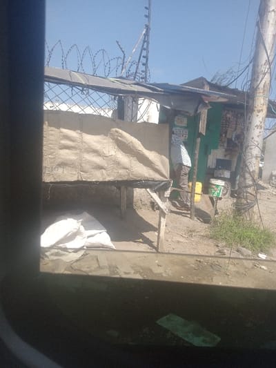 The image depicts an outdoor scene in Ganjoni/Shimanzi ward, Kenya, under a clear blue sky. In the foreground, the lower portion of the image is obscured by a dark, reflective surface, suggesting observation from behind glass. The scene features an informal commercial setup on unpaved ground.
To the left, a makeshift elevated counter or stall is constructed from raw wooden posts, supporting a platform covered with a light brown, possibly burlap or woven sack material. This material drapes down, forming a barrier. Beneath this structure, several white sacks are stacked on the dirt ground.
To the right of the makeshift stall, a single individual stands with their back partially turned towards the camera, facing a green-painted structure. This individual wears a light-colored, possibly striped long-sleeved shirt and dark trousers. The green structure appears to be a small, informal shop or dwelling, with various small items, including packets and what may be merchandise, affixed to or displayed on its exterior. A yellow jerrycan rests on the ground near this structure.
Further to the right, a weathered utility pole with numerous entangled wires extends upwards. The ground throughout the visible area is predominantly dirt, with scattered small patches of green vegetation. In the background, behind the structures, a metal fence topped with coiled razor wire is visible, along with a distinct metal ladder-like structure or antenna extending vertically against the sky.