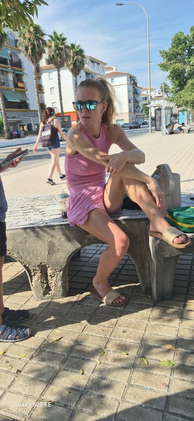 This media file shows a woman sitting on a concrete structure outdoors. She is wearing a pink tank top and pink shorts, with light-colored sandals on her feet. She has sunglasses on and her hair is pulled back. Her legs are crossed, and she is resting her arms on her legs. In the background, there are palm trees, buildings, and a street. Another person, a woman wearing a dark top and denim shorts, is walking in the background. The ground is paved with stone tiles. The scene suggests a sunny day, possibly in the afternoon, given the shadows. The location appears to be a public area, possibly a promenade or a street in a warm climate, consistent with Torre Del Mar, Spain. There are some fallen leaves on the ground. The image is captured in a portrait orientation, and the text "Galaxy S25 FE" is visible in the bottom right corner.