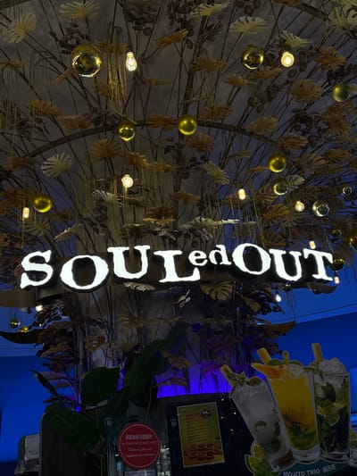 This image shows the interior of a SOULedOUT restaurant, possibly in Genting Highlands, Malaysia, given the sign's multilingual customer service inquiry in Chinese, Hindi, Arabic, and Malay. The vertical "SOULedOUT" sign is the focal point, illuminated against a backdrop of decorative branches adorned with golden ornaments and small light bulbs. A "MOJITO TRIO" advertisement features a beverage display behind the sign. Below it is a dimly-lit area with a menu board labeled "DIMSUMM 精致点心" and a sign indicating customer service availability in multiple languages.