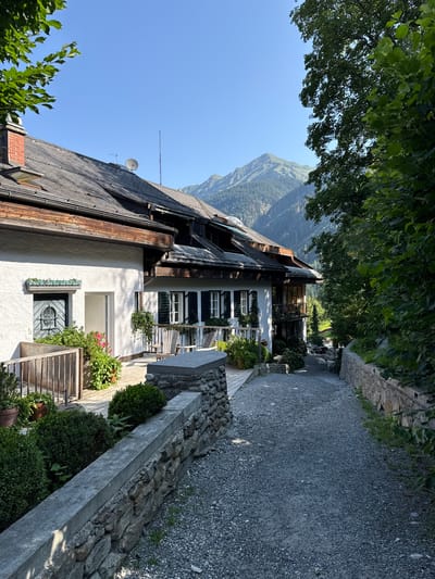 This is a photograph of a picturesque pathway leading to a white building with a dark tiled roof in Bad Gastein, Austria. The building features green shutters on its windows and a balcony. A stone wall lines the right side of the path, which is covered in gravel. Lush green trees and plants surround the scene, and a majestic mountain is visible in the background under a clear blue sky. The scene appears tranquil and bathed in natural sunlight.