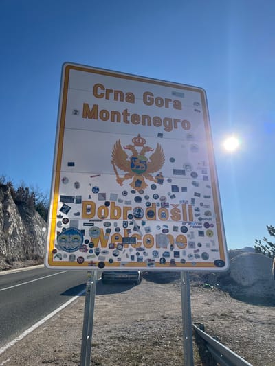 A large, rectangular white and yellow border sign, identifying entry into "Crna Gora Montenegro" with the Montenegrin coat of arms and the greeting "Dobrodošli Welcome," stands prominently at a roadside in Klobuk, Bosnia and Herzegovina. The sign's lower two-thirds are extensively covered with numerous small, multi-colored stickers. It is supported by two grey metal posts. To the left of the sign, an asphalt road with a solid white line runs alongside a rocky hillside with sparse, bare vegetation under a clear blue sky. To the right, a gravel and dirt shoulder features a partially visible light-colored car (license plate "DG BK 769") behind the sign's lower section, a metal guardrail, and scattered large rocks with sparse dry vegetation. The bright sun is visible in the upper right quadrant of the sky, causing lens flare. No individuals are visible.