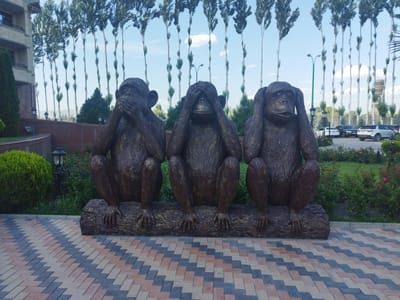 The image shows a statue of three monkeys sitting on a log, with their hands covering their eyes, ears, and mouth, respectively, symbolizing "see no evil, hear no evil, speak no evil." The statue is placed in front of a building with tall trees lining the background. The foreground features a patterned brick walkway and some greenery. The location is identified as Kenbulun, Kyrgyzstan, with a bright sky and clouds visible. Several cars are parked in the background.