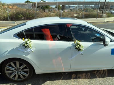 The image shows the side of a white car decorated for a wedding. Two bouquets of white and green flowers are attached to the rear and front door handles with white ribbon. The car's windows are tinted, and through the rear window, red wedding decorations are visible, possibly seat covers or ribbons. There are also some stickers on the inside of the rear window, including what looks like a shoe and a phone.
The car is driving on a road, indicated by the yellow line on the asphalt. In the background, there is a concrete overpass or bridge under construction, with construction fencing and dry vegetation along the roadside. The lighting suggests it is daytime with bright sunlight, casting shadows on the car and the road. The overall scene appears to be in an urban or semi-urban environment, possibly on the outskirts of a town or city. The presence of wedding decorations implies a celebratory event is underway. A blue sticker with white text, partially visible on the rear passenger door, reads "Eldar".