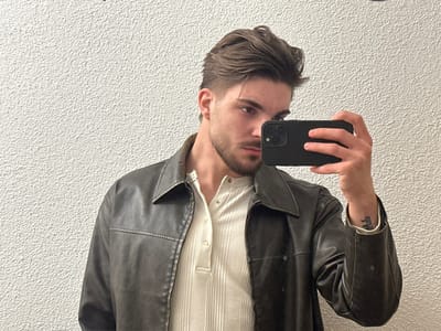 An individual is depicted in an indoor location in Volketswil, Switzerland. The subject is a male, appearing to be in his twenties or early thirties, with medium-brown hair styled upwards and to the right, a trimmed beard, and mustache. He is wearing a dark, possibly brown, collared jacket with a zipper, over a light-colored, ribbed Henley-style shirt with a visible button placket. His right hand holds a black smartphone with a prominent camera array, angled towards the viewer, consistent with capturing a mirror reflection. A small tattoo is visible on the subject's left inner forearm. The background consists of a uniformly lit, light-colored wall with a rough, textured finish.