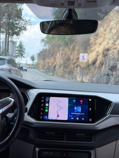The image captures an interior view from a vehicle, looking forward through the windshield. The perspective places the car's dashboard and infotainment system prominently in the lower and right portions of the frame.

The central feature is a large infotainment screen displaying a split interface. The lower section shows Google Maps, indicating a route with the destination "Lago Esmeralda" and a car icon. The top section features a music player widget, showing "SUPERESTRELLA" by "Aitana" as the currently playing song. Visible text on the screen includes "3:50," "4G," and app icons for "Voz" (voice commands), Spotify, and other functions.

Part of the steering wheel is visible in the bottom left, showing some controls and the number "25." To the right of the infotainment screen, air vents and a hazard light button are seen, with the text "PASSENGER AIR BAG" printed above.

Through the windshield, a road is visible, with other vehicles suggesting the car is in traffic or stopped. In the distance, a white rectangular sign with a red circle and a crossed-out letter "E" clearly indicates a "no parking" or "no stopping" zone. The background shows a varied landscape: dry, grassy hills or mountains are visible to the right, while trees and what appear to be distant buildings are seen to the left. The sky is bright and clear, indicating a sunny day.

The car's rear-view mirror is visible in the upper center of the frame, reflecting a person's head with light-colored hair, likely the driver or a front-seat passenger. The context of "Lago Esmeralda" and Atizapán de Zaragoza, Mexico, suggests this scene is captured within that region.