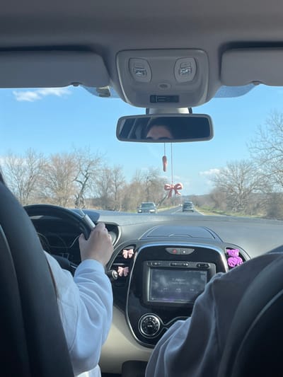 The scene depicts the interior of a vehicle, looking forward through the windshield onto a rural road near the city of Viden, Bulgaria, under a clear blue sky. A person, identified as the driver, is partially visible on the left, wearing a white long-sleeved top and holding the steering wheel with their right hand. Their eyes are reflected in the rearview mirror. Another person, a passenger, is partially visible on the right.
The car's dashboard features a central entertainment screen displaying text "S. Agapao" and "Nikos Vertis." Air vents on the left side of the dashboard are adorned with two pink bow-shaped decorations, while an air vent on the right side holds a purple bear-shaped air freshener. A small pink ballet shoe and a pink bow ornament hang from the rearview mirror. The overhead console contains lights and a passenger airbag indicator.
Outside the vehicle, a two-lane asphalt road stretches into the distance. Bare trees line both sides of the road, suggesting a late winter or early spring setting. Two cars are visible ahead on the road, traveling in the same direction. The surrounding landscape includes gentle hills under the clear sky.