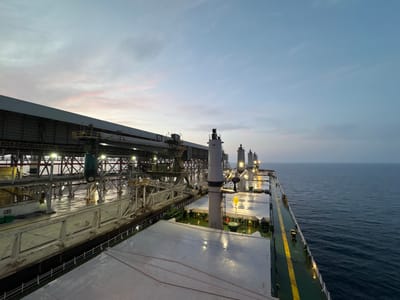 The image captures a view from the deck of a cargo ship docked at the port of Mers El Hadjadj, Algeria. The scene is set at dusk, with a pale blue sky streaked with faint pink clouds. On the left, a large, industrial loading facility with a long, covered structure dominates the waterfront. This facility features multiple conveyor belts, cranes, and support structures, illuminated by artificial lights. The wet surface of the dock suggests recent rain or sea spray.
The cargo ship occupies the foreground and extends into the right side of the frame. Its decks are covered with large, metallic hatches, some of which are reflecting the lights from the port. Several tall, cylindrical structures, likely part of the ship's equipment or cargo handling apparatus, rise from the deck. A bright yellow crane arm is visible, possibly in operation or undergoing maintenance. The dark, calm waters of the sea stretch out to the right, meeting the horizon under the fading twilight. No people are explicitly visible in the frame. The overall impression is one of industrial activity at the close of day.