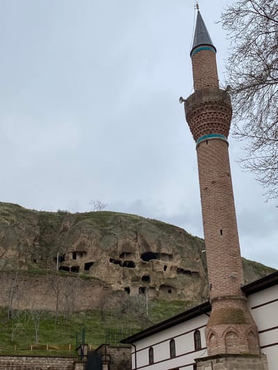 The image shows a tall, brick minaret of a mosque, likely located in Konya, Turkey. The minaret features a distinctive cylindrical structure with bands of ornamentation, including a section with what appears to be openwork or decorative brickwork just below the pointed, cone-shaped roof. The sky is overcast. In the background is a rocky cliff face with several dark cave openings. The area around the cliff appears to be grassy. The architectural style and setting suggest a historical site. Part of a building is visible next to the minaret. Bare tree branches are in the foreground.