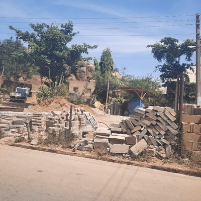 The image captures an outdoor scene, likely a construction material storage area or an informal work site, under a clear blue sky during daylight hours. The main subjects are numerous stacks of light-colored bricks or pavers, with large piles dominating the right side of the frame and smaller stacks extending into the mid-ground. Adjacent to these materials are mounds of dirt or sand.

In the mid-ground, towards the left, a rudimentary structure with a wooden frame and a blue tarp covering is visible, possibly serving as a makeshift shelter or storage space. Further into the background, a building constructed from light-colored bricks or rocks can be seen, partially obscured by green trees and shrubs that are abundant throughout the scene. Prominent large rock formations (boulders) rise behind this building. A white utility vehicle, possibly a small truck, is parked in the lower-left background.

On the far right, a section of a paved road is visible, suggesting the area is adjacent to a public thoroughfare. Overhead, in the upper left portion of the image, a wooden utility pole stands, with multiple power lines stretching horizontally across the clear sky.

The scene depicts no direct human activity, but the presence of construction materials implies ongoing or planned building or landscaping work. The bright sunlight and lack of clouds indicate good, clear weather. No discernible text is visible anywhere in the image. The location is Gerin Gada, Nigeria.