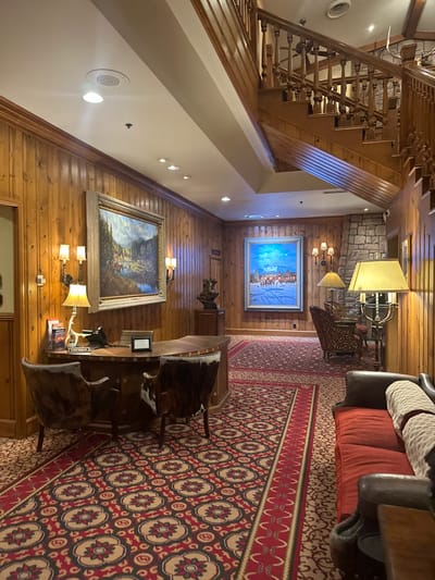 A warm hotel lobby with wood paneling and a patterned rug. Two large landscape paintings hang on the wall, illuminated by sconces. A wooden desk with cowhide-backed chairs sits on the rug. Stairs ascend on the left side of the room. A fireplace with stone detail and lamps is visible. A sofa with red cushions is partially seen on the right. The overall impression is of rustic elegance.