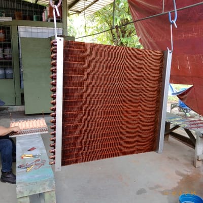 The image shows a large, rectangular, copper-colored cooling coil, suspended from a metal hook in a workshop in Kecamatan Curug, Indonesia, located at coordinates -6.266 latitude and 106.549 longitude. The coil is made of a series of thin, corrugated metal strips, arranged in a vertical pattern. The coil is resting on a concrete floor. There is a wooden table in the foreground, partially obscured by the coil. A man is partially visible on the left side of the image, sitting on the table and holding a metal object. The man is wearing a blue shirt and black pants. In the background, there is a blue tarp and a red tarp hanging from the ceiling. The workshop walls are made of cinder blocks, and there are several unidentified objects in the background. The image is taken from a low angle. A date and time stamp is visible in the lower right corner of the image.