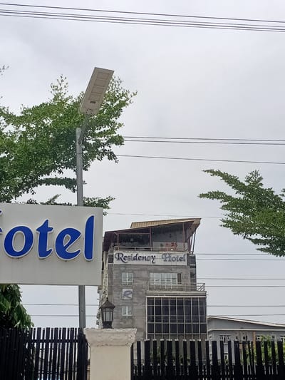 The image presents an outdoor daytime scene featuring architectural structures and lush green foliage under an overcast sky. In the immediate foreground, the partial word "otel" in large blue letters on a white sign is visible, likely part of "Hotel" or "Motel." This sign is partially obscured by abundant green tree branches that also dominate the upper left and right portions of the frame.

A gray solar-powered street light extends upwards on the left side of the image, emerging from the tree canopy. Several dark power lines stretch horizontally across the pale, cloudy sky.

The central focus is a multi-story building identified by a sign on its upper facade as "Residency Hotel" in elegant script. The building appears to be constructed of concrete, with some sections showing a raw finish, potentially indicating ongoing construction or renovation. A large "R" logo is visible on one of the lower floors. The top of the building features a distinctive sloped roof structure, possibly covering a terrace. Below the main hotel sign, there are various windows and a section with a large, grid-patterned glass facade on a lower level.

A black fence with vertical slats runs across the middle ground, partially obscuring the lower parts of the buildings. A white decorative pillar topped with a dark lantern stands as part of this fence structure. To the right and behind the main hotel building, a lower, grey-colored building with a dark roof is visible.

The scene depicts a static view, devoid of people or specific events. The overcast sky suggests a cloudy day, creating soft, diffused lighting. The combination of modern building elements, green trees, and the overall urban landscape provides a visual cue for a developed area like Eputu Town, Nigeria.