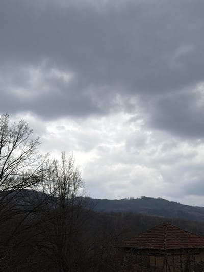 The image depicts an overcast rural landscape in Iamna, Bulgaria. The upper two-thirds of the frame are dominated by dark grey, dense cloud cover, with a brighter, lighter grey area of clouds visible towards the lower center of the sky. In the foreground and midground, the dark silhouettes of multiple leafless deciduous trees are prominent on the left and extend across the center, their intricate bare branches obscuring portions of the view. The lower right quadrant of the image shows the upper section of a building, identifiable by its reddish-brown tiled roof and a segment of a dark, textured wall beneath it, suggestive of aged construction. In the background, a series of dark, undulating hills covered in dense vegetation recedes, culminating in a faint cluster of structures or trees visible atop the furthest ridge. No individuals or overt actions are depicted.