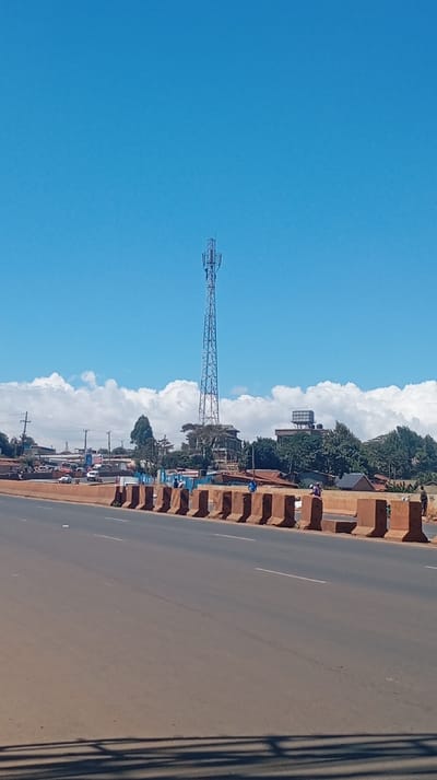 A tall, blue-gray communication tower dominates the skyline under a clear, blue sky dotted with white clouds. A wide, reddish-brown road stretches across the foreground, divided by a row of concrete barriers. Beyond the barriers, there's a mix of trees, buildings, and other structures, indicating an urban or suburban area. People stand near the barriers and along the roadside. A water tower is visible behind the houses. Vehicles are parked along the road.