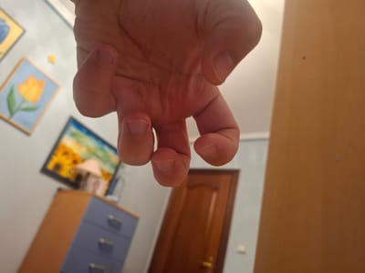A hand is centered in the foreground, obscuring most of the background. The background includes a brown door, a blue and brown dresser, and several framed paintings on a white wall. The photograph was likely taken indoors, in Xirivella, Spain. The exact location in Xirivella cannot be determined from the image.