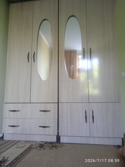 This is a medium shot, eye-level, indoor photograph of a light-colored wooden wardrobe with two sections. Each section has two cabinet doors on top and two drawers on the bottom. The top doors on both sections feature oval mirrors. The handles on the doors and drawers are dark and elongated. The wardrobe is placed on a patterned carpet. The background shows a light green wall. The date and time stamp "2026/7/17 08:59" is visible in the bottom right corner. The scene appears to be a domestic setting, likely a bedroom, in Лесхоз, Kyrgyzstan.