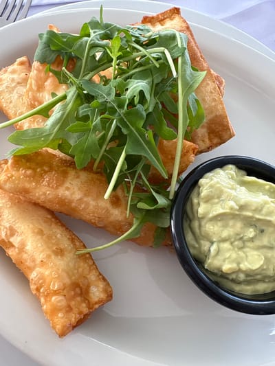 A white plate holds an appetizer of fried spring rolls topped with fresh arugula and a side of avocado dip. The spring rolls are golden brown and appear to be filled with a savory mixture. The arugula is a vibrant green and is artfully arranged on top of the rolls, adding a fresh contrast. The avocado dip is creamy and pale green, served in a small black bowl. The setting is likely a restaurant in La Punta, Peru, given the presentation and style of the dish, and the focus on fresh ingredients.