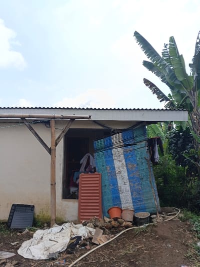 A small, run-down building with a corrugated metal roof is the focal point. A makeshift wooden support structure is visible on the left. Items such as buckets, blue and white striped sheets, and a red plastic panel are stacked against the building's wall. Discarded materials and debris are scattered on the ground in front, with a white cloth covering some of it. Dense foliage, including banana trees, grows to the right, contributing to the somewhat unkempt appearance of the property. The sky is partly cloudy.