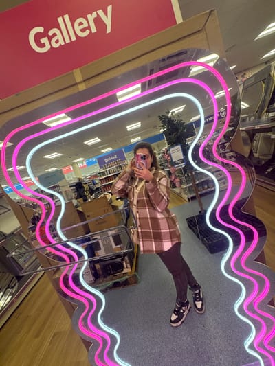 The image captures a reflection in a wavy, heart-shaped mirror with pink and white neon lights around the edge. The reflection shows a person holding a phone, presumably taking the photo. The surroundings reflected in the mirror appear to be a retail store, potentially a "Gallery" as indicated on the side of the mirror's packaging, which suggests the store is located in Portadown, United Kingdom. The mirror is positioned on a display stand. There are shelves stocked with merchandise and other boxes or packaging visible in the reflection. The floor in the reflection appears to be carpeted or has a solid surface, and the lighting in the store is bright and typical of a retail environment.