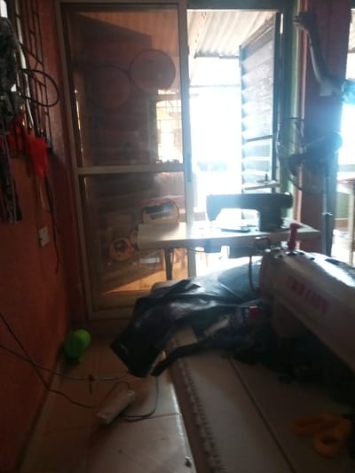 The image shows a dimly lit room, likely a workshop or a home sewing area, in Agbara, Nigeria. Two sewing machines are visible. The machine in the foreground is a beige "TWO LION" brand, partially obscured by dark fabric. The second sewing machine, a black industrial model, sits on a table positioned in front of a large glass door that opens to the outside. The door itself has a corrugated metal roof structure visible through it, suggesting an outdoor or semi-outdoor space beyond.

On the left side of the image, the wall is painted an orangish-red color. A metal grate or rack is attached to the wall, with some red and black fabric items hanging from it. A green plastic bottle and electrical cords are on the floor near the wall.

Inside the room, beyond the glass door, stacked circular items are visible against a wall, possibly for storage or display. The overall impression is a functional workspace, perhaps a bit cluttered, with natural light filtering in from the open doorway. The time of day is indeterminate, but the lighting suggests it could be daytime, with the bright light from outside creating a contrast with the interior. No people are visible in the image.