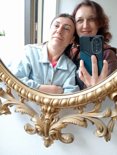 This media file is a portrait of two women taken in Kuopio, Finland. The image appears to be a mirror selfie, with the two women posing in front of a mirror with an ornate, gold-colored frame. Both women are looking at the camera, with one woman on the left leaning her head slightly on the shoulder of the woman on the right.

The woman on the left has dark brown hair pulled back and is wearing a light blue denim jacket over a pink top. The woman on the right has curly, reddish-brown hair and is wearing a dark hoodie. She is holding a smartphone in her right hand, with the screen visible and oriented towards the mirror to capture the reflection. The phone appears to have a dual-camera system.

The background is a plain white wall, with a glimpse of a window frame to the left. The lighting suggests it is daytime, possibly indoors with natural light from the window. The overall mood seems casual and friendly. There is no visible text in the image.