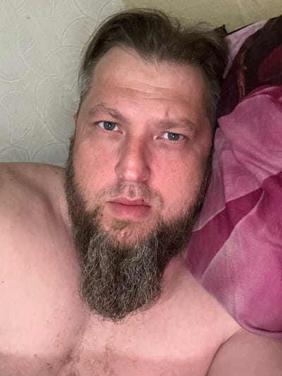 A man with a beard and blue eyes is lying in bed, looking at the camera. He is wearing a pink blanket. The room is dimly lit and the wall behind him has a textured pattern. The image appears to be a selfie taken indoors.